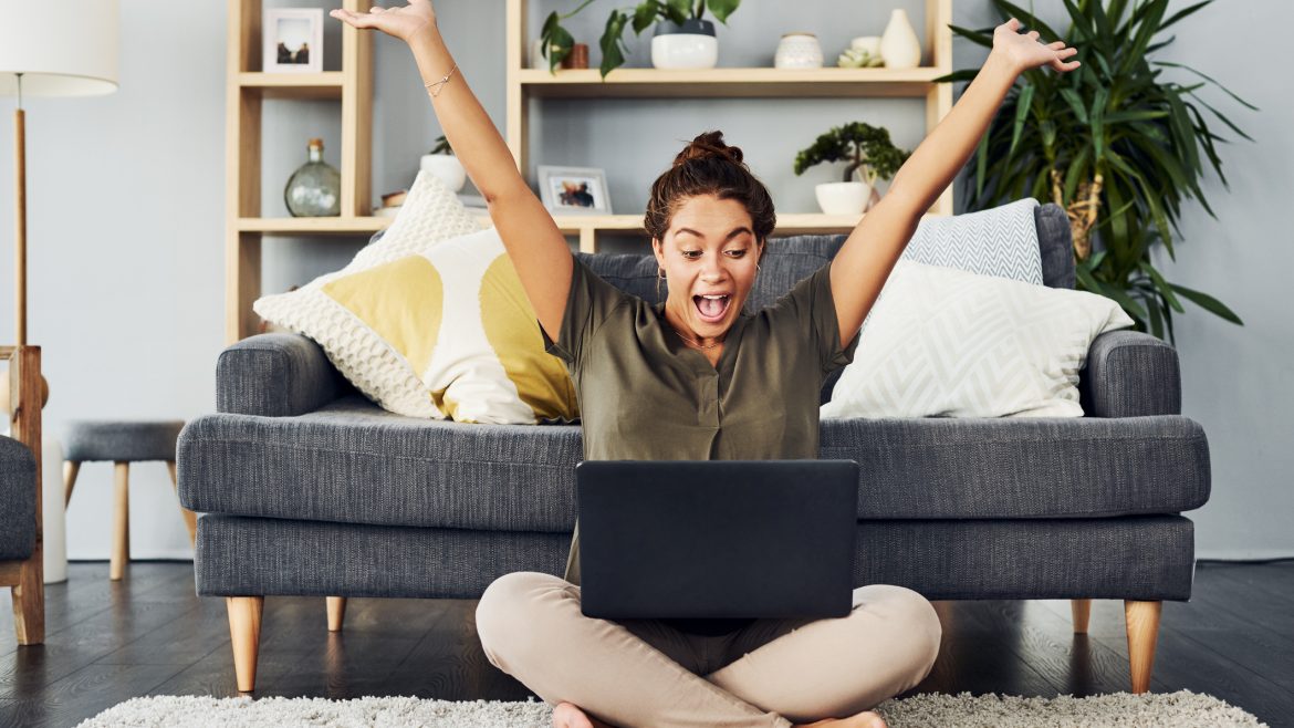 Shot of a young woman cheering while using a laptop on the living room floor at home