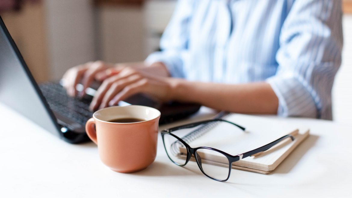 A woman typing on a laptop. A notebook and pen are beside her on the desk, with a pair of eyeglasses and a cup of coffee.