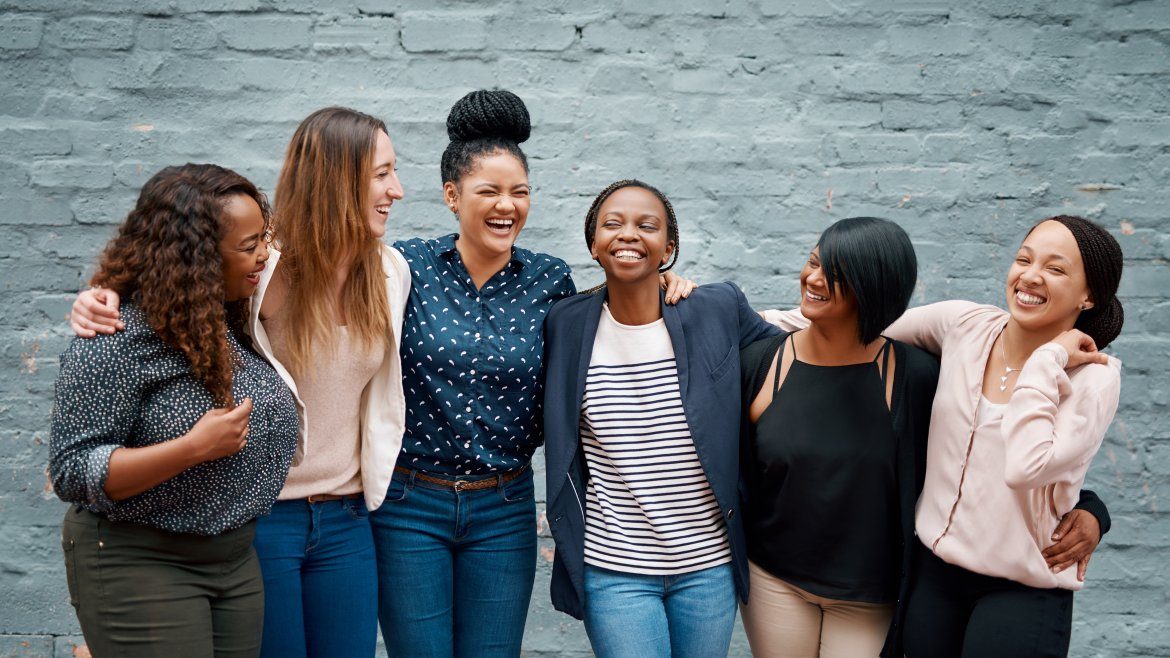 A group of young women stand together in front of a wall. They have their arms around each other and are laughing.