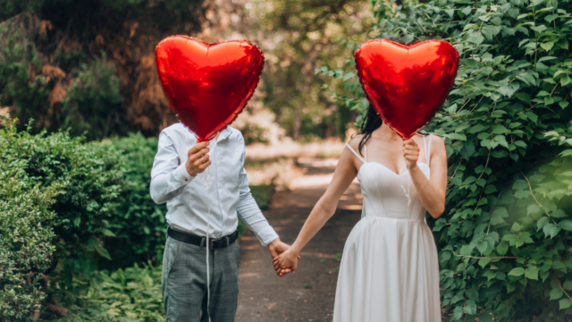 Two people stand in a garden holding heart shaped red balloons over their faces
