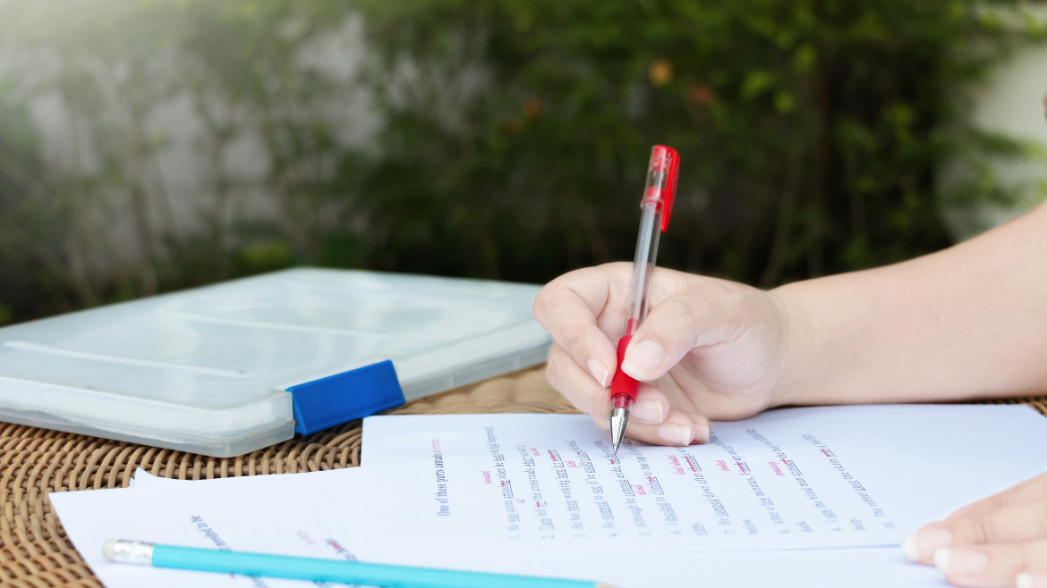 Paper being edited with a red pen. An arm rests on the page on the right side