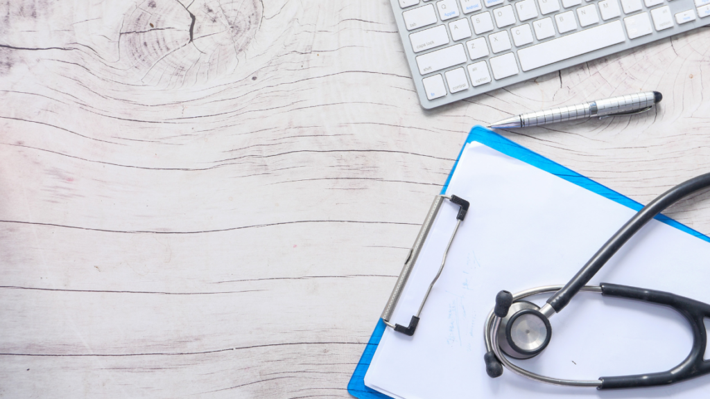 A blue clipboard with a stethoscope resting on top sits on a white wooden desk.  A computer keyboard sits in the top right corner. 