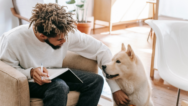 A man is sitting in a chair with a notebook in his lap and is petting a white dog