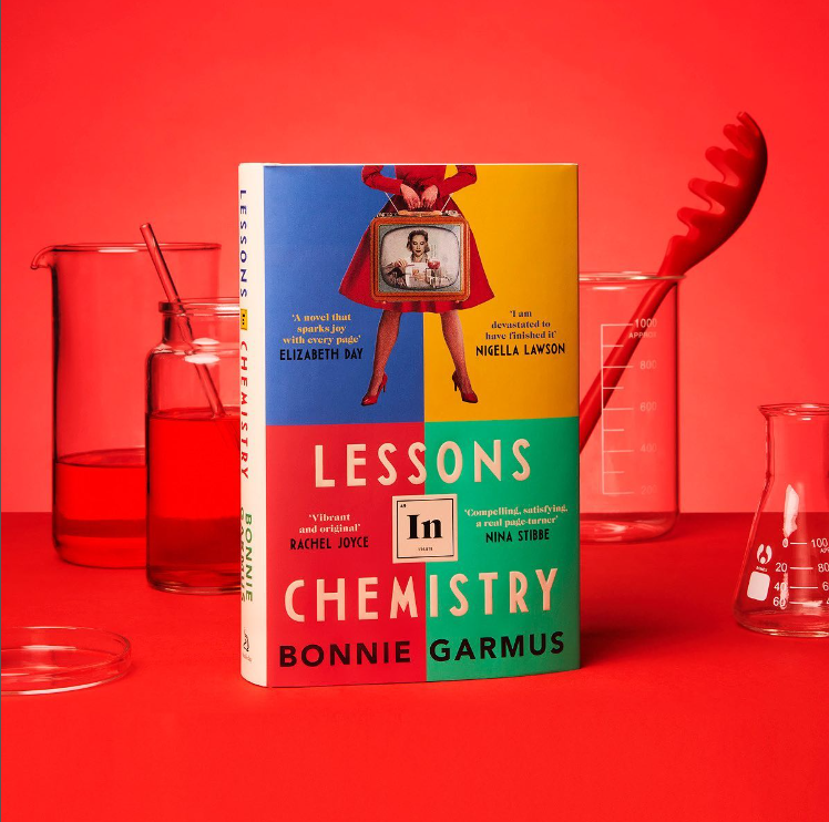 A copy of Bonnie Garmus' book Lessons in Chemistry on a red backdrop with beakers behind it. 