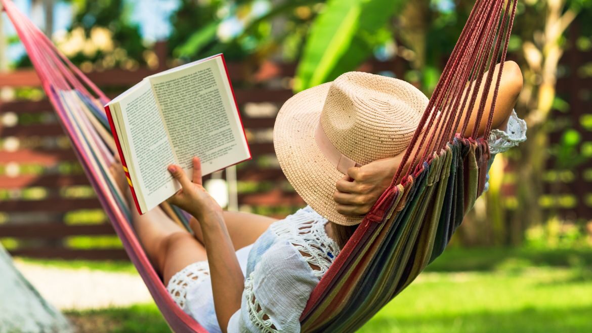 A woman in a straw hat reads a book in a hammock on a sunny day.