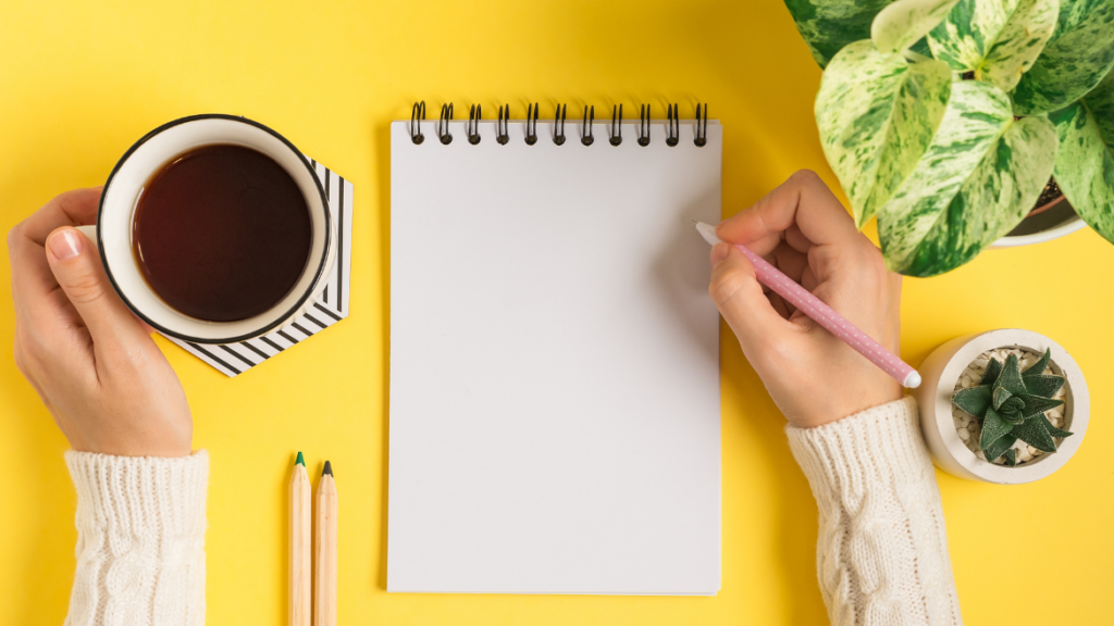 A white notebook on a yellow background with two hands on either side.  The left hand holds a coffee and the right, a pencil