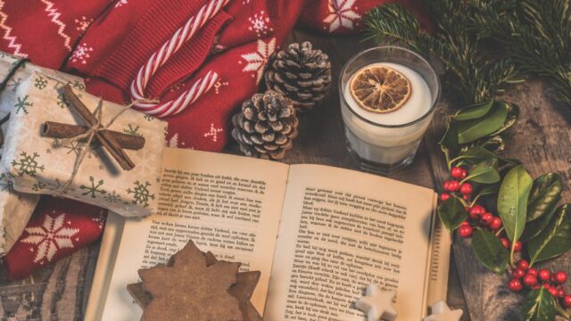 A selection of festive items on a table, including a Christmas jumper, a candy cane, a book, pine cones, a glass of eggnog and some holly leaves.