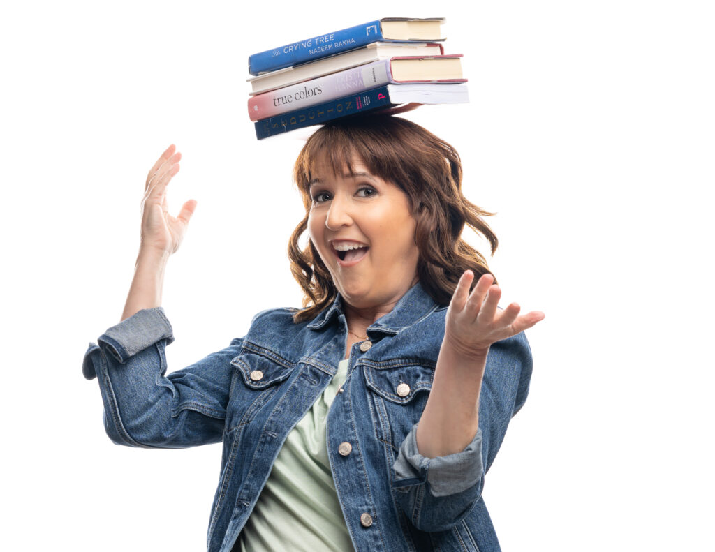 Author Kate MacGuire, a white woman with brown hair, stands in front of a white backdrop and balances books on her head.
