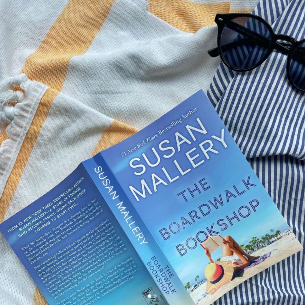 Susan Mallery's novel, The Boardwalk Bookshop, sits on a white and yellow striped beach towel next to a pair of sunglasses and a blue and white striped pant leg