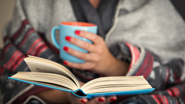 A women with red nails is wrapped in a red, plaid blanket and holding a blue book and a blue mug.