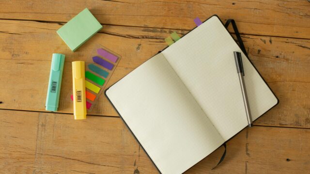 An open notebook on a desk. A pen, highlighters and post sticks around the notebook.