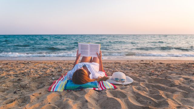 A woman lays on the beach reading a book on a blue towel.