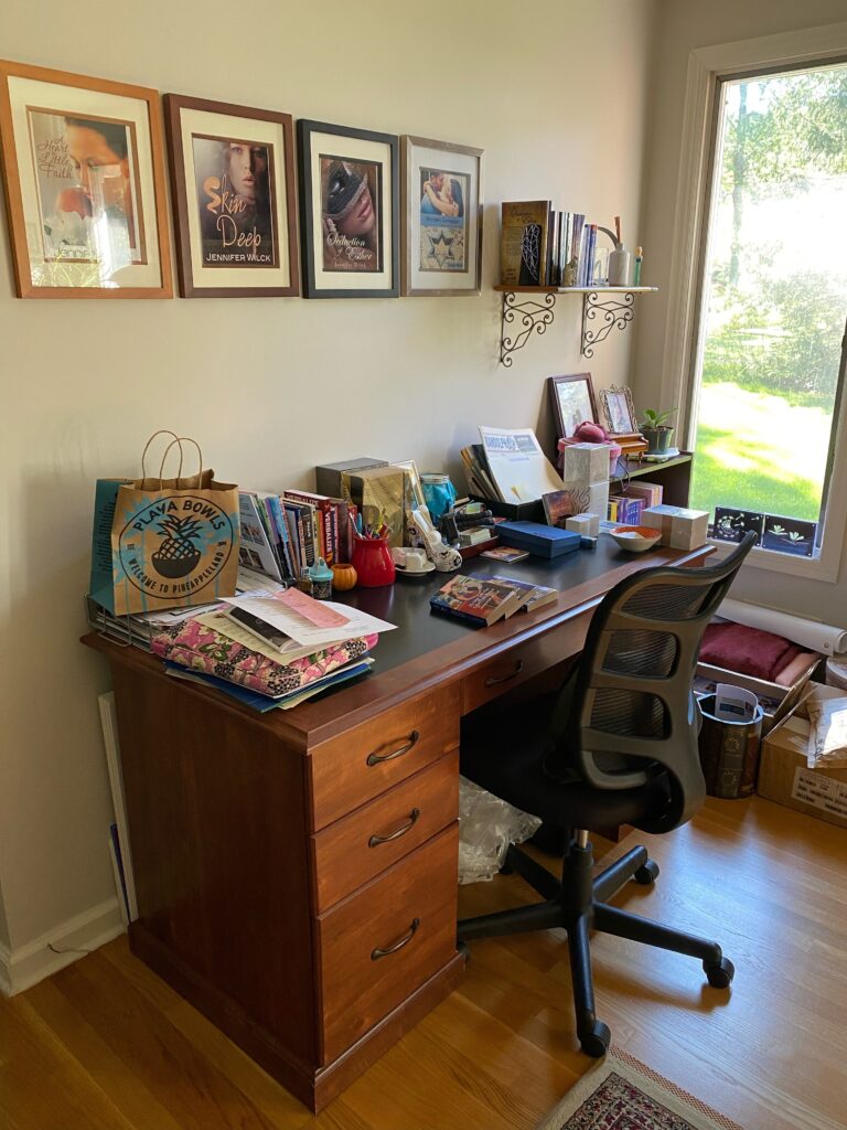 A brown wood desk is covered in books along the left wall of a bright room.  The window on the right casts light over the room. 