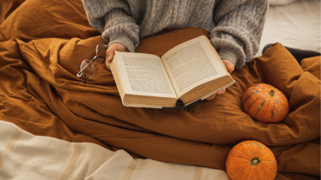 A woman in a grey sweater sits under an orange blanket with an open book in her lap