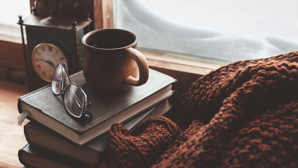 In front of a snowy window, a book and a pair of glasses sits on a stack of books next to an orange blanket