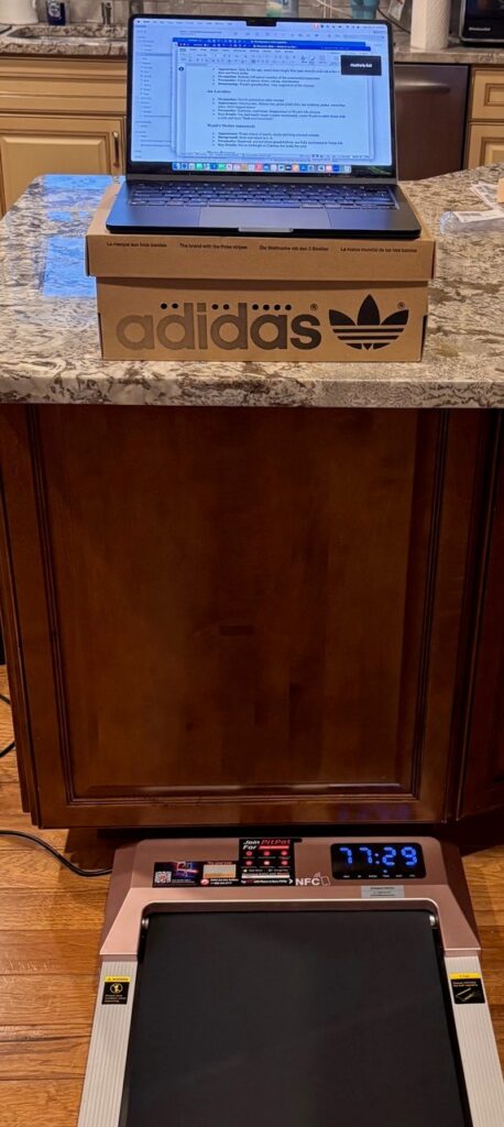 A laptop sits on an adidas shoe box on a kitchen counter. A walking pad sits on the floor below